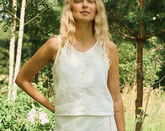 Puede incluir: Una mujer con un conjunto de lino blanco, compuesto por un top sin mangas con botones y una falda cruzada. El conjunto se encuentra en un entorno de exuberante vegetación, con árboles y flores, creando una estética natural.