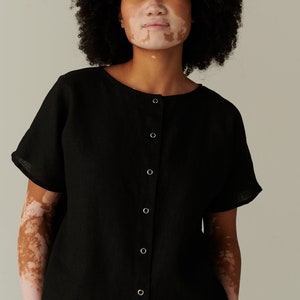 Pu&ograve; includere: Una camicia nera in lino con bottoni e maniche corte. La camicia ha un collo rotondo e una patta con bottoni sul davanti.