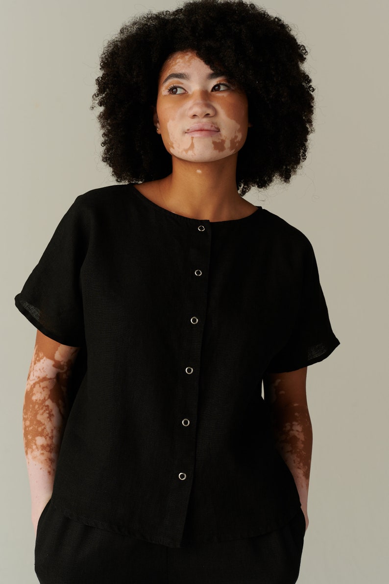 Pu&ograve; includere: Una camicia nera in lino con bottoni e maniche corte. La camicia ha un collo rotondo e una patta con bottoni sul davanti.