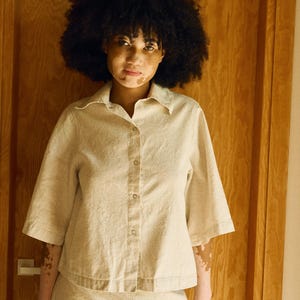 May include: A beige button-down shirt with short sleeves and a matching skirt. The outfit is made of a textured fabric. The model has dark curly hair and skin with vitiligo. The background is a wooden door.