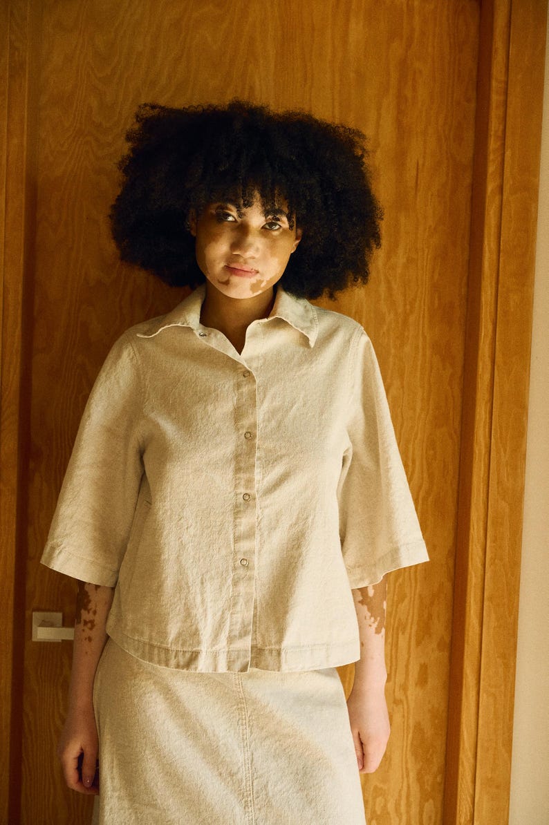 May include: A beige button-down shirt with short sleeves and a matching skirt. The outfit is made of a textured fabric. The model has dark curly hair and skin with vitiligo. The background is a wooden door.