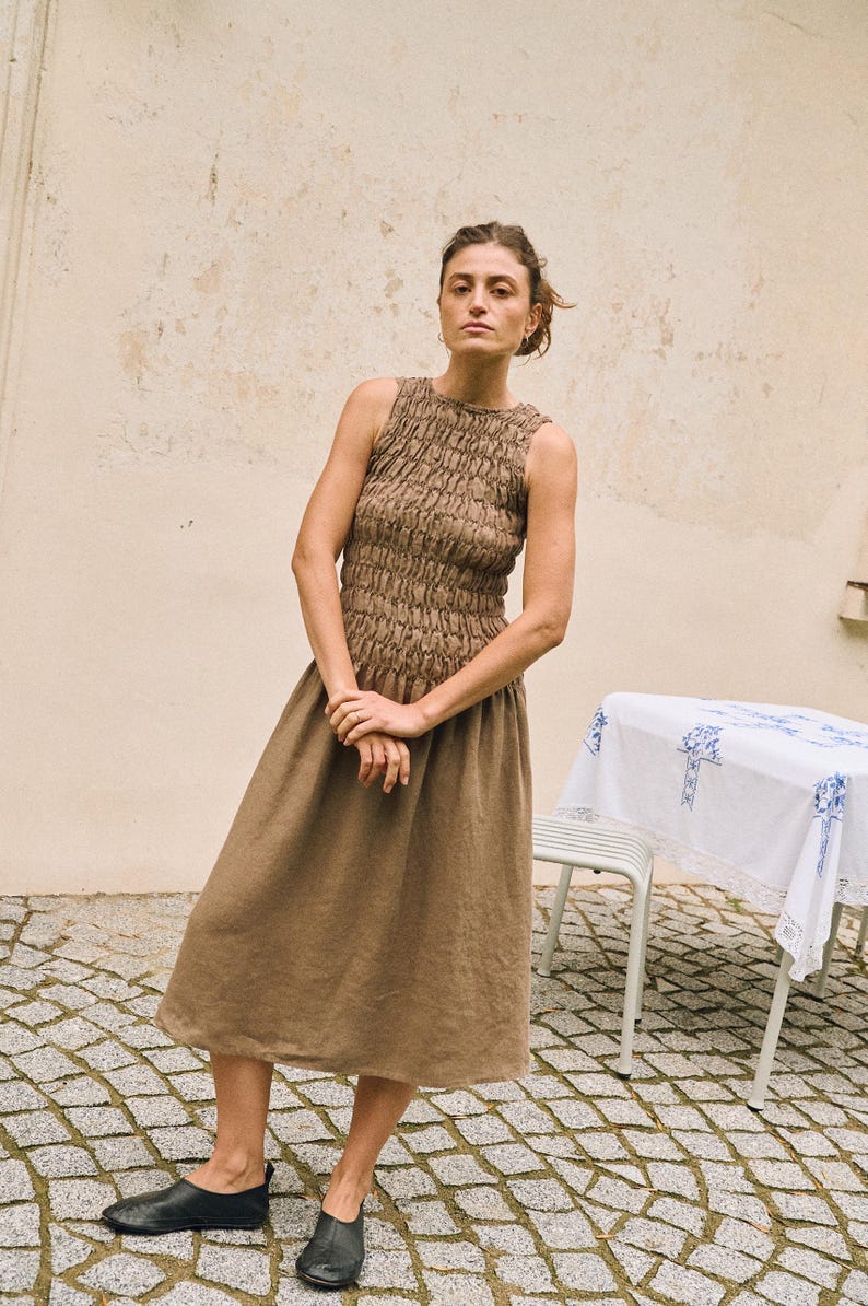 May include: A woman wearing a sleeveless, brown dress with a textured bodice and a flowing skirt. She is wearing black shoes. The dress is knee-length. The background is a neutral-toned wall and a stone patio.