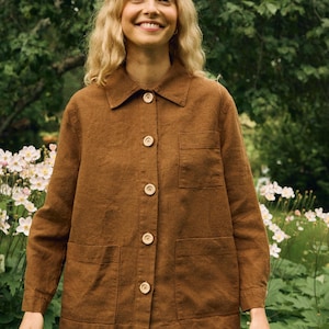 Puede incluir: Chaqueta de lino marrón con cuello, cierre de botones y dos bolsillos delanteros. La chaqueta se combina con pantalones cortos verde oliva. El fondo presenta vegetación y flores blancas.