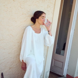 May include: A person wearing a white linen top and pants, drinking from a clear glass. The top has a square neckline with lace trim and long, puffy sleeves. The person is standing outside near a textured wall.