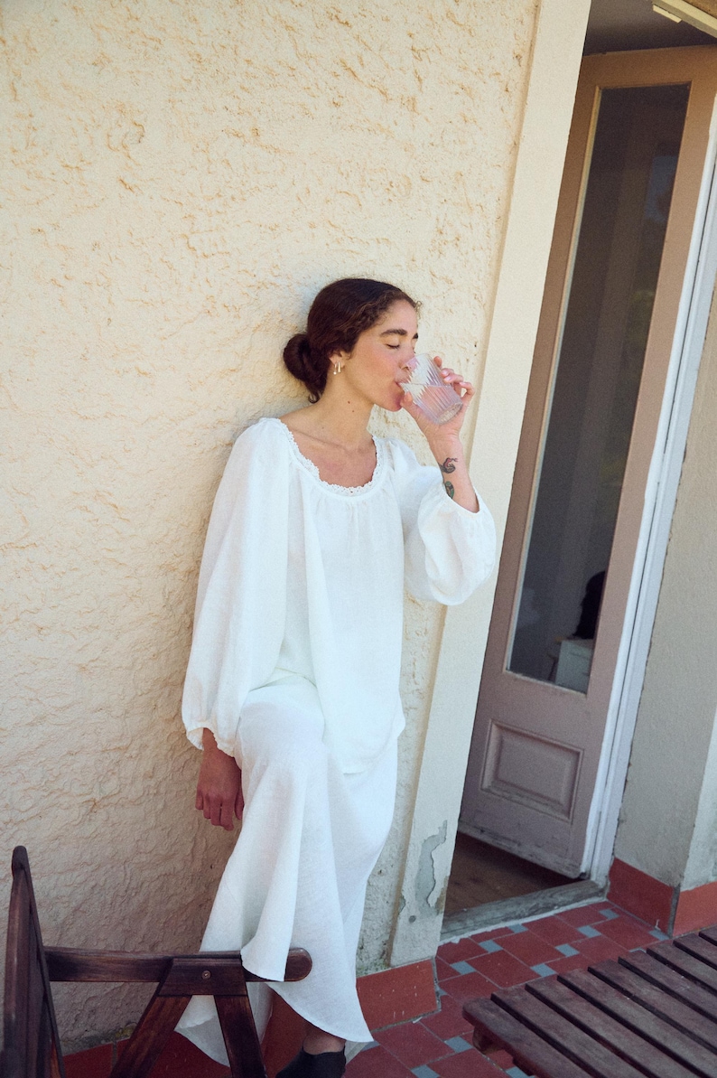 May include: A person wearing a white linen top and pants, drinking from a clear glass. The top has a square neckline with lace trim and long, puffy sleeves. The person is standing outside near a textured wall.
