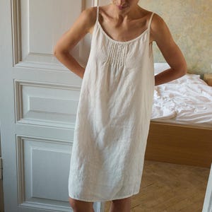 Puede incluir: Un vestido de slip de lino blanco con tirantes finos y un detalle fruncido en el pecho.