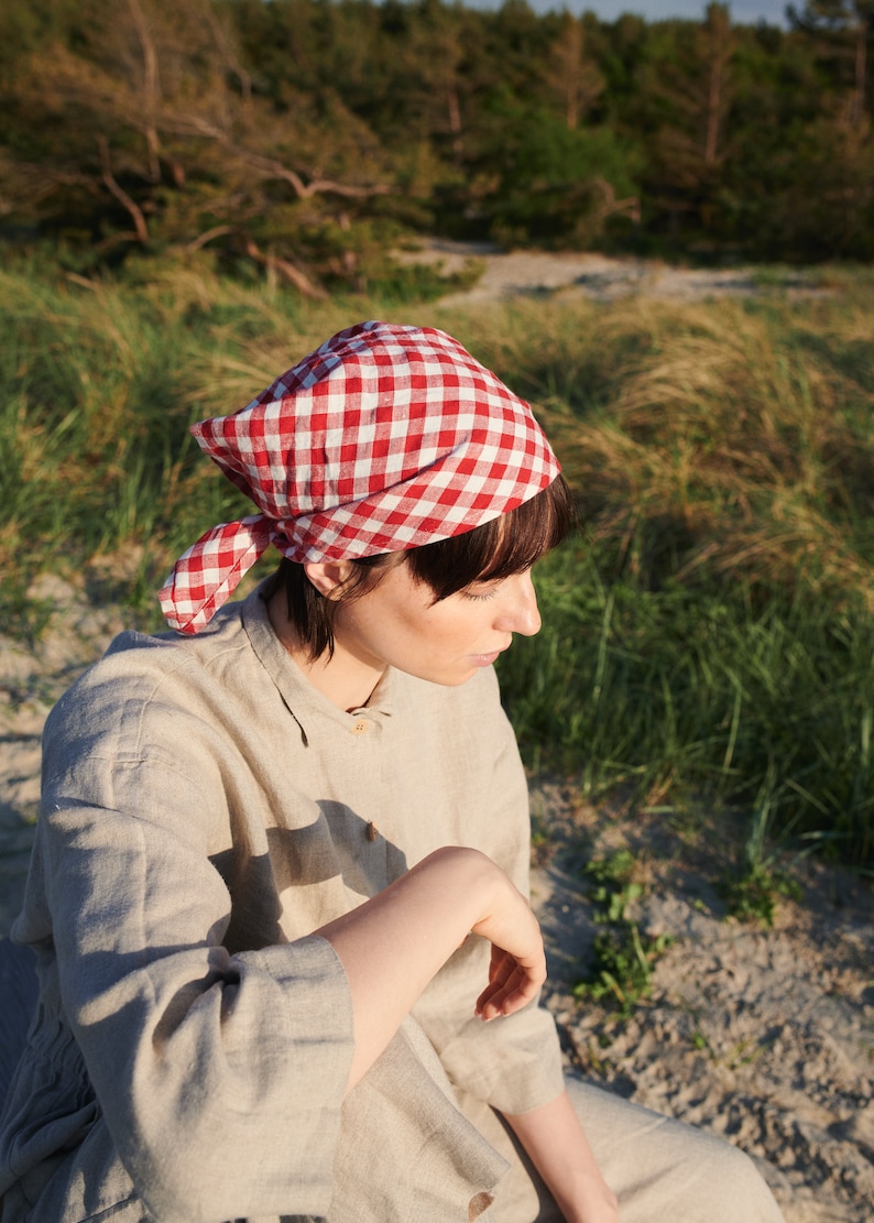 May include: A person wearing a red and white gingham bandana and a light beige linen shirt. The person is sitting in a grassy area.