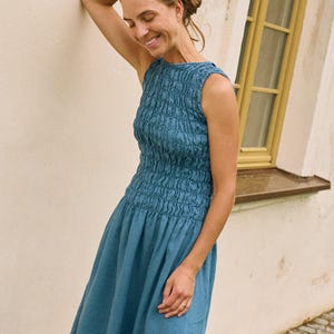 May include: A sleeveless, blue dress with a textured bodice and a flowing skirt. The dress is knee-length and appears to be made of linen or a similar lightweight fabric. The woman is smiling and leaning against a wall.