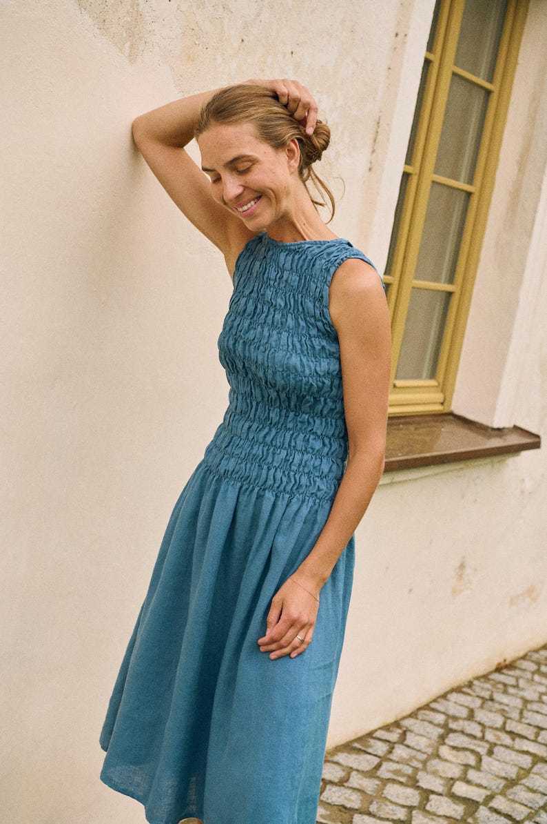 May include: A sleeveless, blue dress with a textured bodice and a flowing skirt. The dress is knee-length and appears to be made of linen or a similar lightweight fabric. The woman is smiling and leaning against a wall.