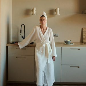 以下が含まれることがあります： 白いワッフル生地のローブに、結ぶベルトが付いています。ローブは、白いタオルを頭に巻いた人が着用しています。その人は、白いキャビネットのあるキッチンに立っています。