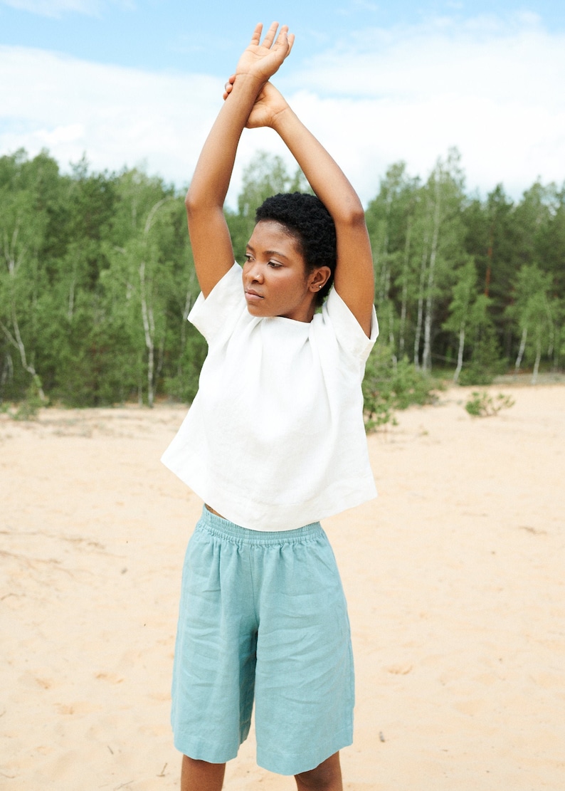 May include: A woman wearing a white linen top and light blue linen shorts stands in a sandy area with trees in the background. She is stretching her arms above her head.