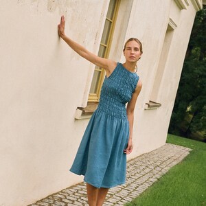 May include: A sleeveless, blue dress with a textured bodice and a flared skirt. The model is wearing black shoes and is leaning against a white wall next to a grassy area. The dress is knee-length.