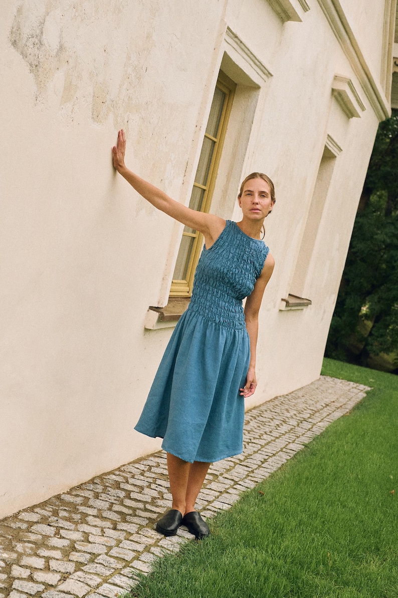 May include: A sleeveless, blue dress with a textured bodice and a flared skirt. The model is wearing black shoes and is leaning against a white wall next to a grassy area. The dress is knee-length.