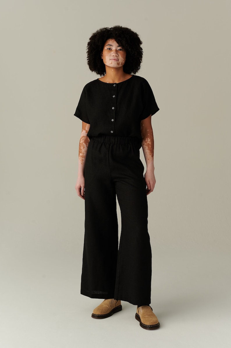 Pu&ograve; includere: Una donna indossa una tuta in lino nero con una parte superiore abbottonata e una vestibilit&agrave; rilassata. Indossa mocassini in camoscio marroni.
