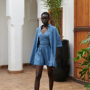 May include: A person wearing a blue linen shirt and matching shorts. The shirt is unbuttoned and the shorts have an elastic waistband. The person is wearing black leather sandals and standing in a courtyard with a palm tree in the background.