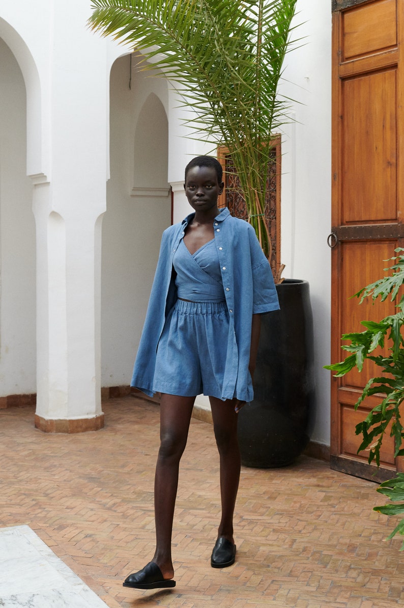 May include: A person wearing a blue linen shirt and matching shorts. The shirt is unbuttoned and the shorts have an elastic waistband. The person is wearing black leather sandals and standing in a courtyard with a palm tree in the background.