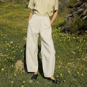 Può includere: Un paio di pantaloni di lino color crema a gamba larga con vita a coulisse. I pantaloni sono indossati da una persona in piedi in un campo di fiori gialli.
