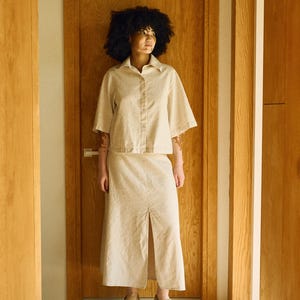 May include: A beige linen shirt and skirt set. The short-sleeved button-down shirt and midi skirt have a relaxed fit. The skirt features a front slit. The person is wearing beige slip-on shoes. The outfit is in a neutral color palette.