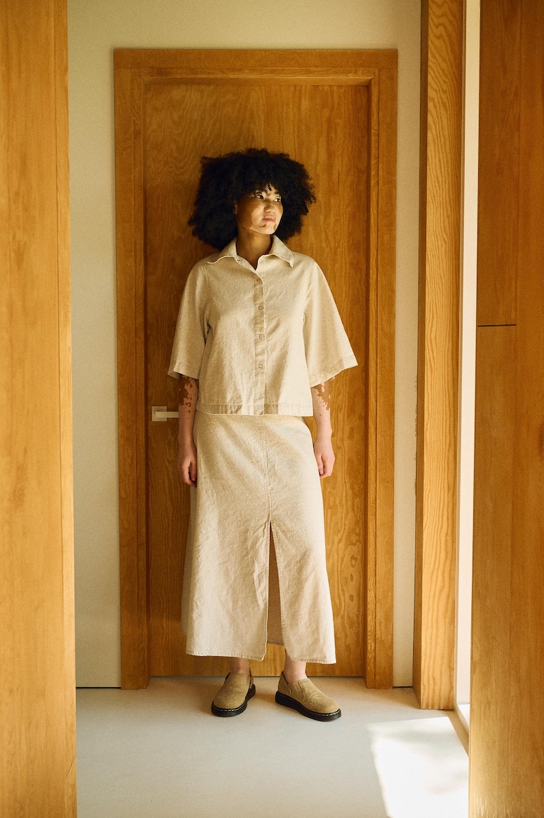May include: A beige linen shirt and skirt set. The short-sleeved button-down shirt and midi skirt have a relaxed fit. The skirt features a front slit. The person is wearing beige slip-on shoes. The outfit is in a neutral color palette.