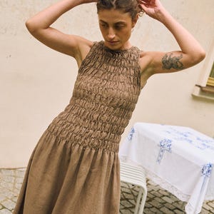 May include: A brown sleeveless dress with a textured bodice and a flowing skirt. The dress has a high neckline and a gathered waist. The model is wearing the dress outdoors. A table with a white tablecloth is in the background.