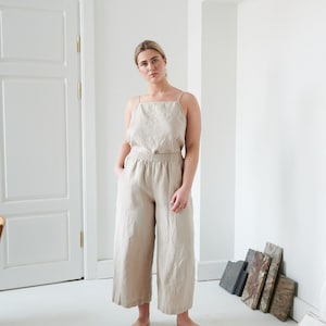 May include: A woman wearing a light beige linen jumpsuit with thin straps. The jumpsuit has a relaxed fit and wide legs. The woman is standing in a white room with a white door in the background.