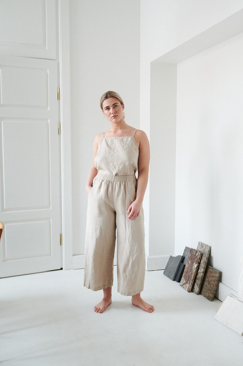 May include: A woman wearing a light beige linen jumpsuit with thin straps. The jumpsuit has a relaxed fit and wide legs. The woman is standing in a white room with a white door in the background.