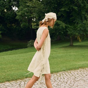 May include: A sleeveless, light yellow dress with tie details, paired with black leather slides and a matching headscarf. The person is walking on a stone path in a park with green grass and trees.