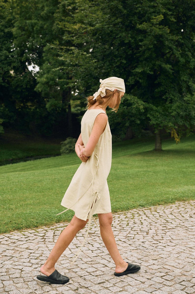 May include: A sleeveless, light yellow dress with tie details, paired with black leather slides and a matching headscarf. The person is walking on a stone path in a park with green grass and trees.