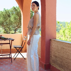 May include: A woman in a sleeveless, checked top and white trousers with decorative stitching, paired with black shoes. The setting is an outdoor patio with terracotta tiles and arched architecture.