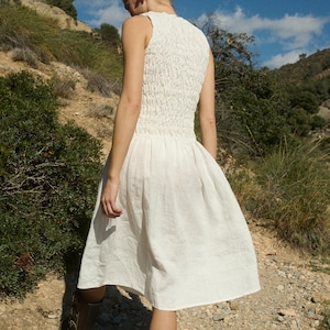 May include: A white sleeveless dress with a gathered bodice and a flowing skirt. The dress is worn by a person walking on a dirt path.