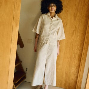 May include: A beige linen two-piece set, consisting of a button-down shirt with short sleeves and a midi skirt with a front slit. The outfit is paired with tan suede slip-on shoes. The model is standing in a doorway.