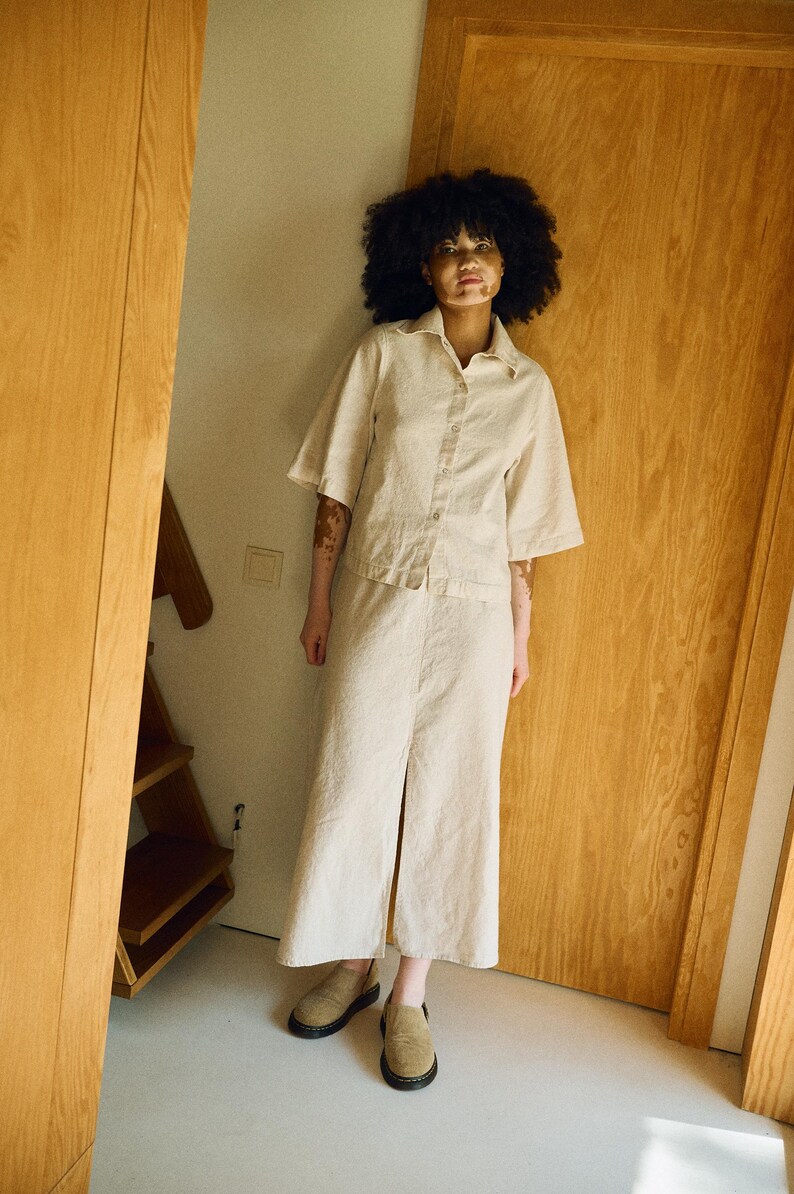 May include: A beige linen two-piece set, consisting of a button-down shirt with short sleeves and a midi skirt with a front slit. The outfit is paired with tan suede slip-on shoes. The model is standing in a doorway.