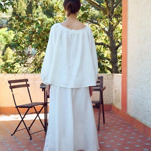 May include: A white linen two-piece outfit, consisting of a long-sleeved top and a flowing skirt. The top has a scoop neckline. The person is standing on a red tile patio with two wooden chairs and a small table. The background includes trees and a building.