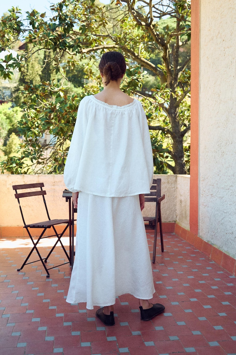 May include: A white linen two-piece outfit, consisting of a long-sleeved top and a flowing skirt. The top has a scoop neckline. The person is standing on a red tile patio with two wooden chairs and a small table. The background includes trees and a building.