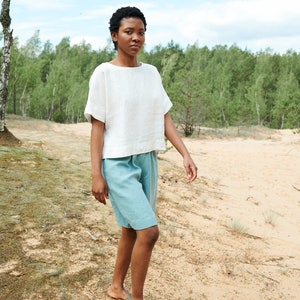 May include: A person wearing a white linen top and light blue linen shorts walks on a sandy path. The person is wearing brown sandals.