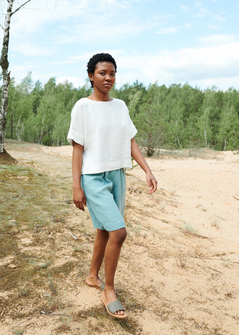 May include: A person wearing a white linen top and light blue linen shorts walks on a sandy path. The person is wearing brown sandals.