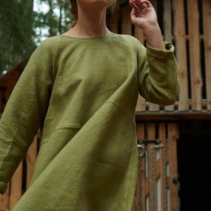 Peut inclure: Une femme portant une robe en lin vert &agrave; manches longues et &agrave; col rond. La robe est ample et a une jupe l&eacute;g&egrave;rement &eacute;vas&eacute;e. Elle se tient devant une structure en bois.
