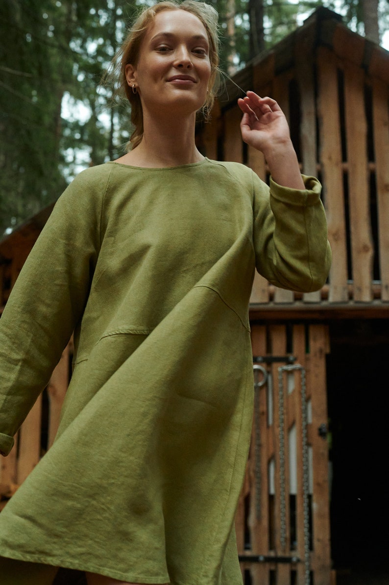 Peut inclure: Une femme portant une robe en lin vert &agrave; manches longues et &agrave; col rond. La robe est ample et a une jupe l&eacute;g&egrave;rement &eacute;vas&eacute;e. Elle se tient devant une structure en bois.