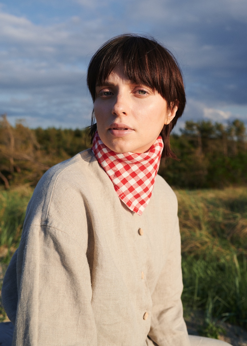 May include: A woman with short brown hair wearing a beige linen shirt with a red and white gingham bandana tied around her neck. She is standing in a field of tall grass.