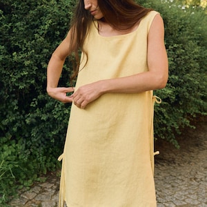 May include: A sleeveless, pale yellow linen dress with a square neckline and side ties. The dress is paired with a matching headscarf with lace trim. The outfit is simple and elegant, perfect for warm weather.