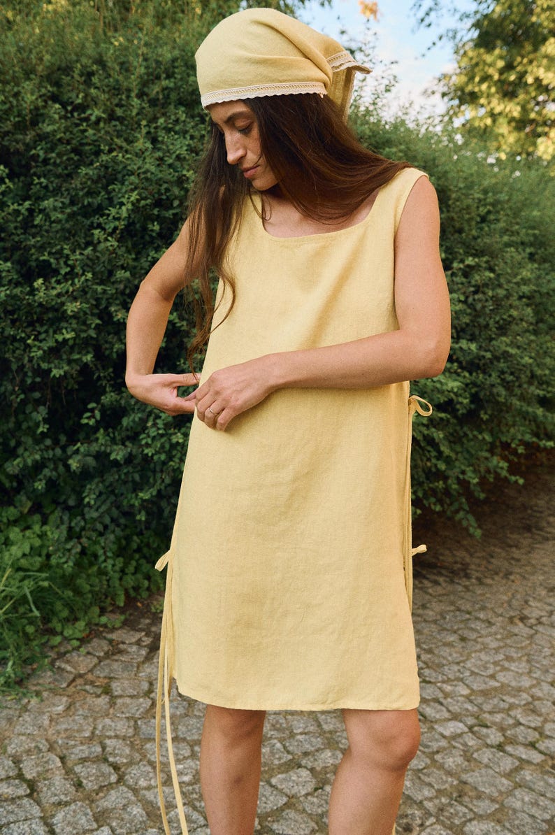 May include: A sleeveless, pale yellow linen dress with a square neckline and side ties. The dress is paired with a matching headscarf with lace trim. The outfit is simple and elegant, perfect for warm weather.
