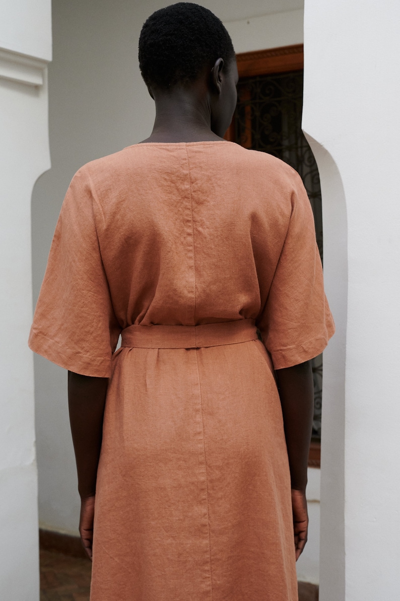Puede incluir: Un vestido de lino terracota con cintura ce&ntilde;ida. El vestido tiene un corte sencillo y relajado y una falda ligeramente acampanada.