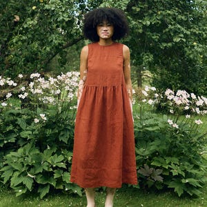 May include: A sleeveless, rust-colored dress with a gathered waist. The dress is knee-length and worn with tan shoes. The background features a lush green garden with white flowers and a large tree.