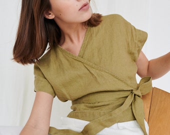 May include: A woman wearing a green linen wrap top with short sleeves and a tie at the waist. She is sitting on a wooden chair and wearing white pants.