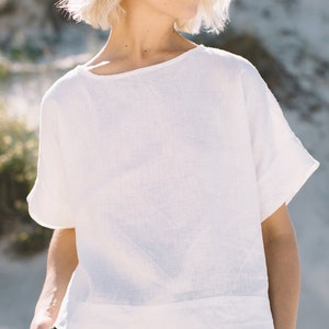 May include: A white linen top with short sleeves and a relaxed fit. The top has a boat neckline and a slightly cropped hem. The model is wearing the top with a dark green linen skirt.