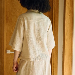 May include: A beige linen shirt and skirt set. The short-sleeved shirt has a button-down front and a small collar. The skirt is a midi length. The outfit is made of a lightweight fabric and is suitable for warm weather.