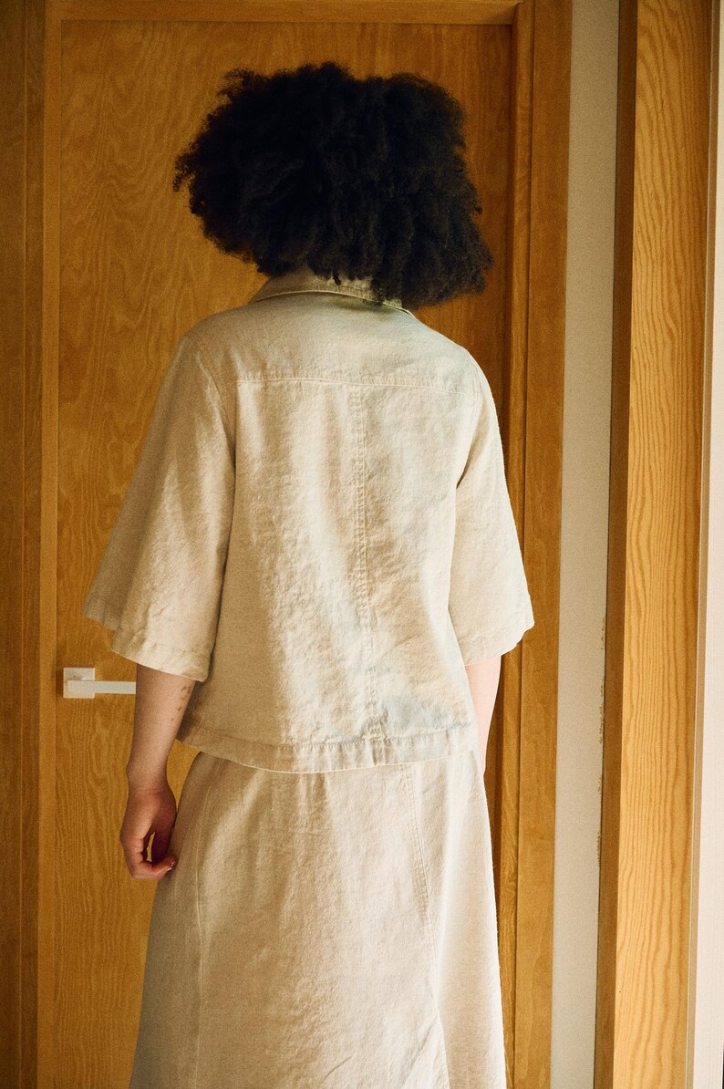 May include: A beige linen shirt and skirt set. The short-sleeved shirt has a button-down front and a small collar. The skirt is a midi length. The outfit is made of a lightweight fabric and is suitable for warm weather.