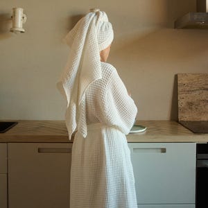 以下が含まれることがあります： 白いワッフル生地のローブを着て、頭にタオルを巻いた人がキッチンに立っています。