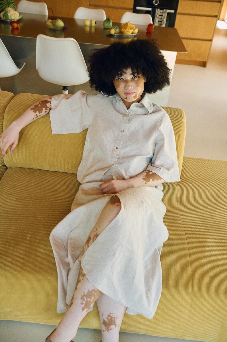 May include: A person wearing a light beige button-down shirt and matching skirt, seated on a yellow corduroy sofa. The outfit has short sleeves and a knee-length skirt. The person has dark curly hair.