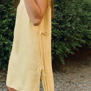 May include: A sleeveless, pale yellow linen dress with side ties. The dress has a simple, flowing design and a knee-length hemline. The side ties add a decorative element. The dress is made of a lightweight fabric, perfect for warm weather.